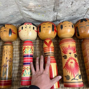 May include: A selection of traditional Japanese Kokeshi dolls. The dolls feature round heads with painted faces and cylindrical bodies. The bodies are adorned with red, yellow, and green floral and geometric patterns. Some dolls have writing on them.
