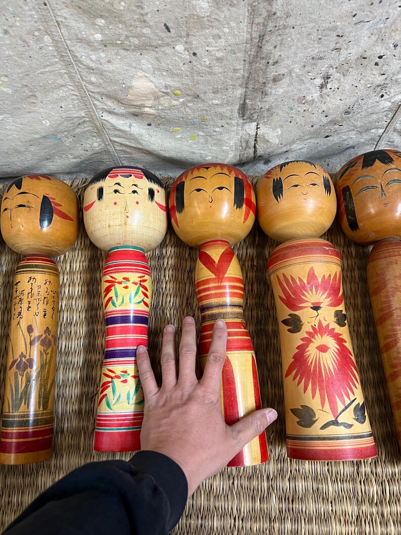 May include: A selection of traditional Japanese Kokeshi dolls. The dolls feature round heads with painted faces and cylindrical bodies. The bodies are adorned with red, yellow, and green floral and geometric patterns. Some dolls have writing on them.
