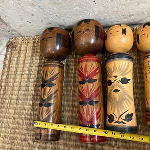 May include: A selection of Japanese Kokeshi dolls. The dolls feature wooden cylindrical bodies and rounded heads. The bodies are adorned with painted floral and geometric patterns in red, black, and brown. The heads have painted facial features and black hair.