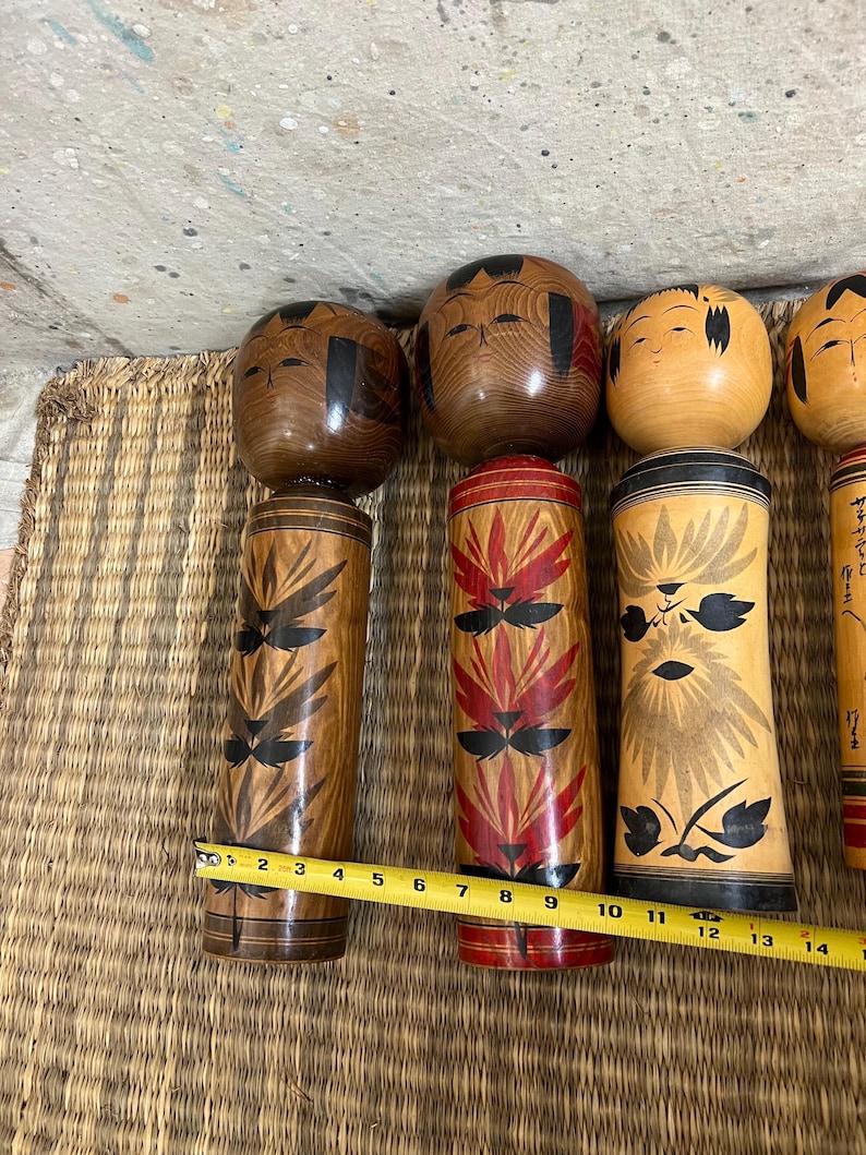 May include: A selection of Japanese Kokeshi dolls. The dolls feature wooden cylindrical bodies and rounded heads. The bodies are adorned with painted floral and geometric patterns in red, black, and brown. The heads have painted facial features and black hair.