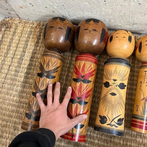 May include: A collection of Japanese Kokeshi dolls. The dolls have cylindrical bodies with painted floral designs in red, black, and brown. The heads are round and painted with simple facial features. The dolls are arranged on a woven mat.