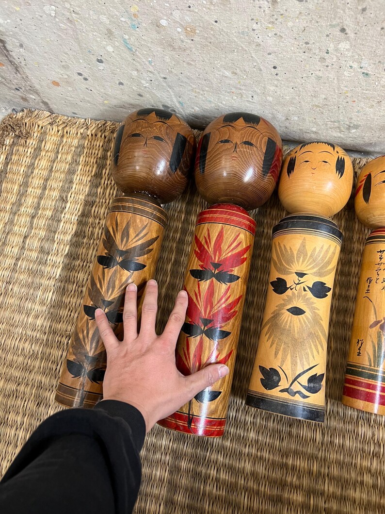 May include: A collection of Japanese Kokeshi dolls. The dolls have cylindrical bodies with painted floral designs in red, black, and brown. The heads are round and painted with simple facial features. The dolls are arranged on a woven mat.