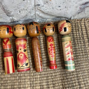 May include: Six wooden Kokeshi dolls are displayed. Each doll has a round head with painted faces and a cylindrical body. The bodies are decorated with red, black, and green floral and geometric patterns.