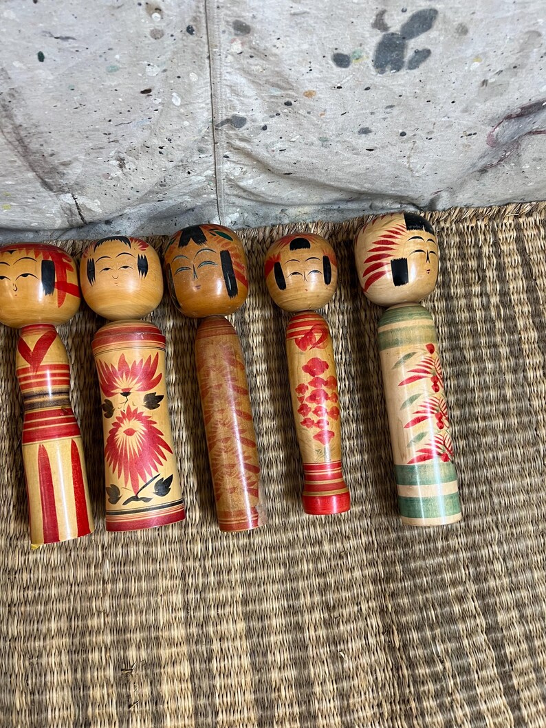 May include: Six wooden Kokeshi dolls are displayed. Each doll has a round head with painted faces and a cylindrical body. The bodies are decorated with red, black, and green floral and geometric patterns.