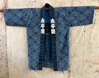 Op de afbeelding: Een blauw en wit Japans jasje met een geometrisch patroon en witte geborduurde karakters. Het jasje heeft een donkerblauwe kraag en een donkerblauwe band.