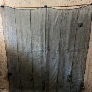 Könnte beinhalten: Ein großes, rechteckiges, halbtransparentes Stoffpaneel in einem dunklen, gedeckten Blaugrau. Das Textil weist vertikale Nähte und sichtbare Flicken aus dunklerem Stoff auf und ist mit zwei schwarzen Metallklammern an einem Holzstab befestigt.