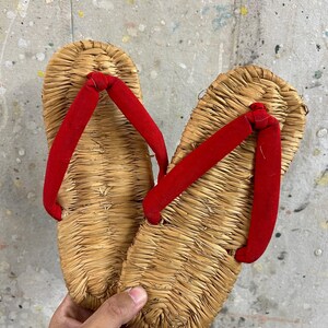 以下が含まれることがあります： 編み込まれた藁のソールと鮮やかな赤色の布製鼻緒が特徴の伝統的な日本のサンダル。この履物は、天然の乾燥植物繊維で作られた平らな楕円形のベースを持ち、中央に赤いストラップが固定されています。