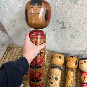 May include: A wooden Kokeshi doll with a round head and cylindrical body. The head is light brown with black hair and red accents. The body is decorated with red and black leaf-like patterns. Other Kokeshi dolls are visible.