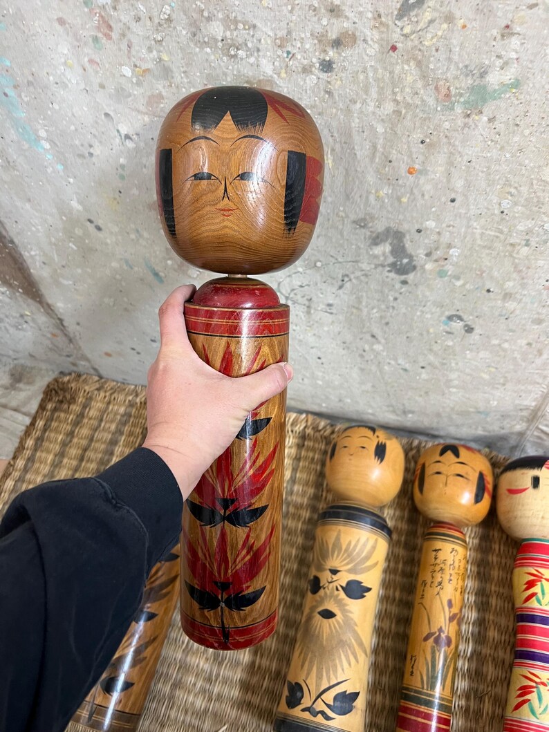 May include: A wooden Kokeshi doll with a round head and cylindrical body. The head is light brown with black hair and red accents. The body is decorated with red and black leaf-like patterns. Other Kokeshi dolls are visible.