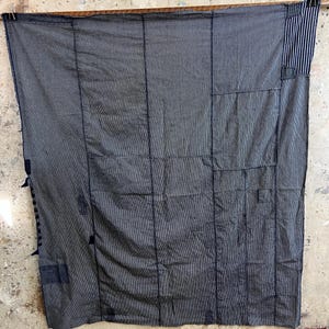 Könnte beinhalten: Ein dunkelgraues Textil mit Patchwork-Design, das vertikale Nähte und rechteckige Patches aufweist. Der Stoff hat ein subtiles Streifenmuster und ist an einer Holzstange aufgehängt. Das Textil hat ein rustikales, handgefertigtes Aussehen.