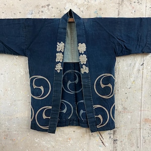 May include: A dark blue indigo-dyed cotton jacket featuring wide sleeves and a front opening. The garment displays light-coloured circular tomoe patterns and Japanese kanji characters printed vertically on the front lapels in a light beige tone.