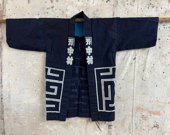May include: Navy blue Japanese kimono-style jacket with white geometric designs and Japanese characters. The jacket has wide sleeves and a blue lining. The garment is hanging on a wooden rod against a neutral background.
