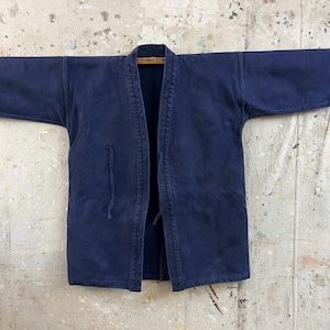 Könnte beinhalten: Eine dunkelmarineblaue Jacke mit Sashiko-Nähten, texturiertem Steppmuster, weiten Ärmeln und offenem Vorderteil mit dünnen Stoffbändern. Das Kleidungsstück hat einen strukturierten Kragen und einen geraden Saum in einem einheitlichen Indigo-Farbton.