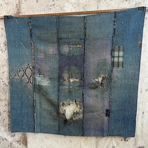 Puede incluir: Un tapiz mural textil azul con un diseño de patchwork. La pieza presenta varios tonos de azul, con costuras visibles y parches de diferentes telas. El textil está colgado de una barra de madera.