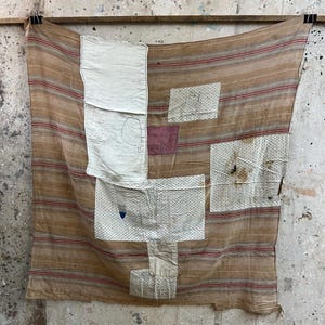May include: A rectangular textile piece featuring a tan and brown striped base with multiple stitched-on fabric patches. The patches include a large white rectangular section, smaller patterned white squares, and a solid dark red square piece.