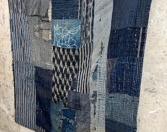 May include: A patchwork textile in shades of blue and grey, with visible stitching and varying fabric patterns. The textile is hung on a wooden rod with a clip. The design includes stripes, floral motifs, and geometric shapes, creating a rustic, handcrafted aesthetic.