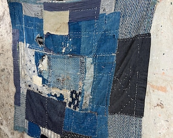 May include: A patchwork textile in shades of blue, indigo, and cream, with visible stitching. The fabric is composed of various textured patches, including stripes and dotted patterns. The textile is suspended from a wooden rod with black clips.