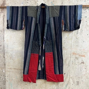 Könnte beinhalten: Vintage Kimono mit Streifen in Marineblau, Grau und Weiß. Das Kimono hat rote Patches am unteren Ende und grüne Patches auf den Schultern. Das Kleidungsstück hängt an einer Holzstange vor einer gefleckten Wand.