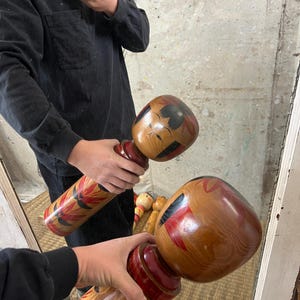 May include: Two wooden Kokeshi dolls are held in front of a mirror. The dolls have a rounded head and cylindrical body. The heads are painted with black and red details. The bodies feature red and floral designs.