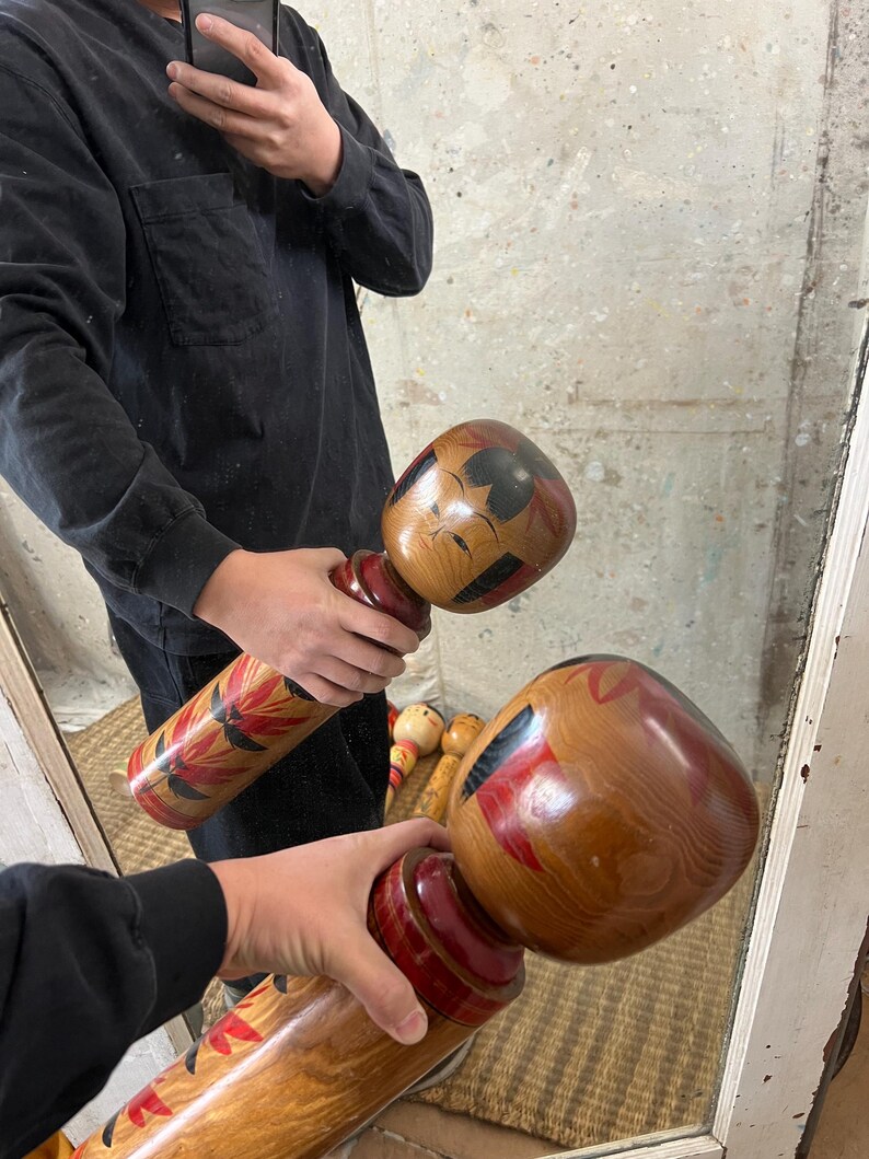 May include: Two wooden Kokeshi dolls are held in front of a mirror. The dolls have a rounded head and cylindrical body. The heads are painted with black and red details. The bodies feature red and floral designs.