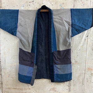 Puede incluir: Una chaqueta estilo kimono de patchwork con un diseño de bloques de color en tonos azul índigo, gris pizarra y negro. La prenda presenta mangas anchas, un cuello estructurado y un forro interior azul oscuro con un sutil patrón de cuadrícula.