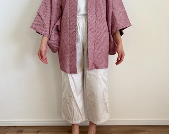 Puede incluir: Chaqueta burdeos estilo kimono con tejido texturizado. La chaqueta tiene mangas anchas y un corte holgado. La persona lleva una camiseta blanca y pantalones blancos. El fondo es una pared blanca lisa y un suelo de madera.