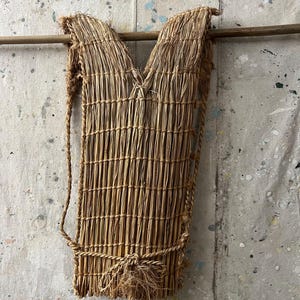 May include: A woven straw bag with a V-shaped top and a rectangular body. The bag has a natural, light brown color and is suspended by a rope. The bag is made of tightly woven straw and has a rustic, handmade appearance.