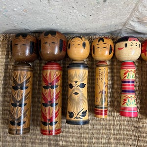 May include: A selection of Japanese Kokeshi dolls. The dolls feature round heads with painted faces and cylindrical bodies. The bodies are adorned with diverse patterns in brown, red, and black. The dolls are displayed on a woven mat.