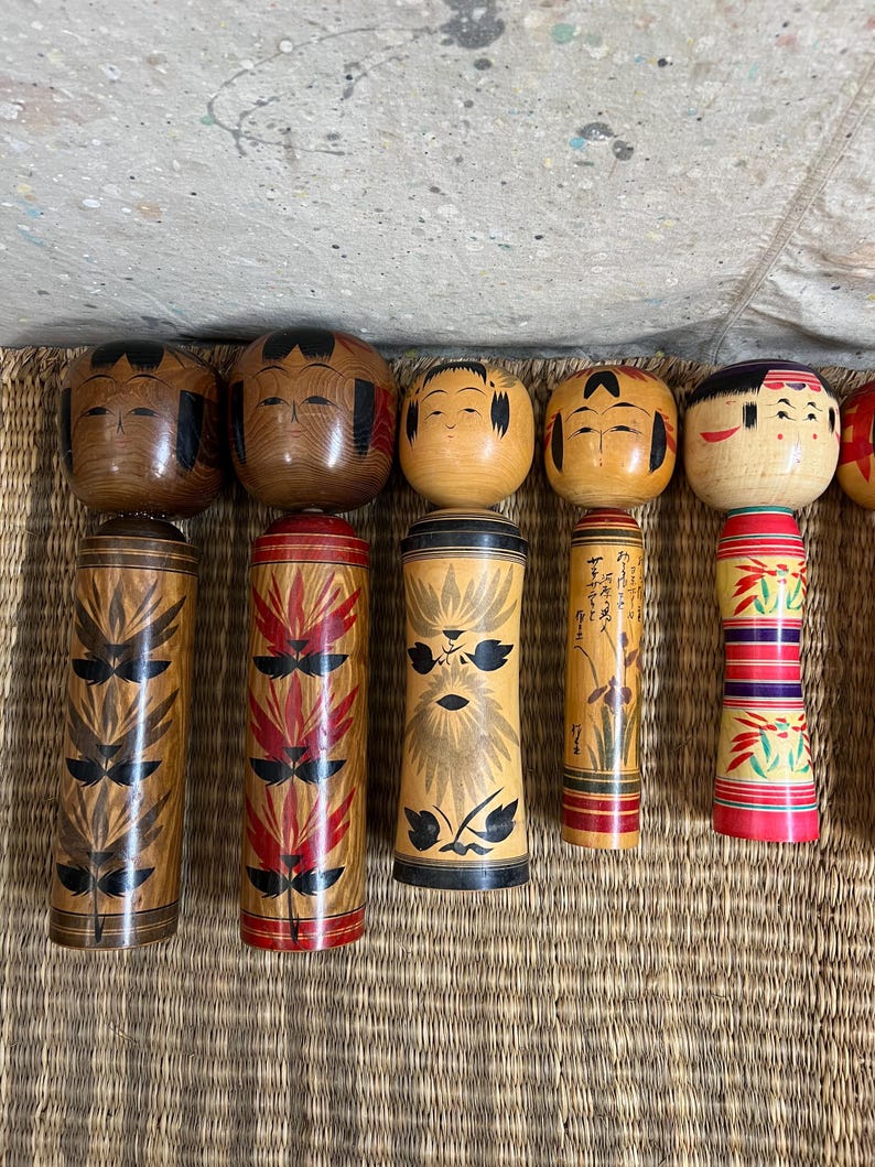 May include: A selection of Japanese Kokeshi dolls. The dolls feature round heads with painted faces and cylindrical bodies. The bodies are adorned with diverse patterns in brown, red, and black. The dolls are displayed on a woven mat.