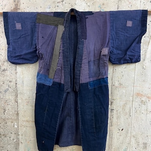 Puede incluir: Una chaqueta índigo de estilo boro con varios parches de tela en tonos azul y azul marino oscuro. La prenda presenta costuras visibles, múltiples reparaciones rectangulares de tela y una construcción en capas con mangas anchas.