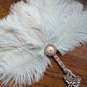 May include: A light blue feather fan with a decorative silver handle. The handle features a pearl and rhinestone embellishment at the top and a floral design at the base. The fan is made of soft, fluffy feathers.