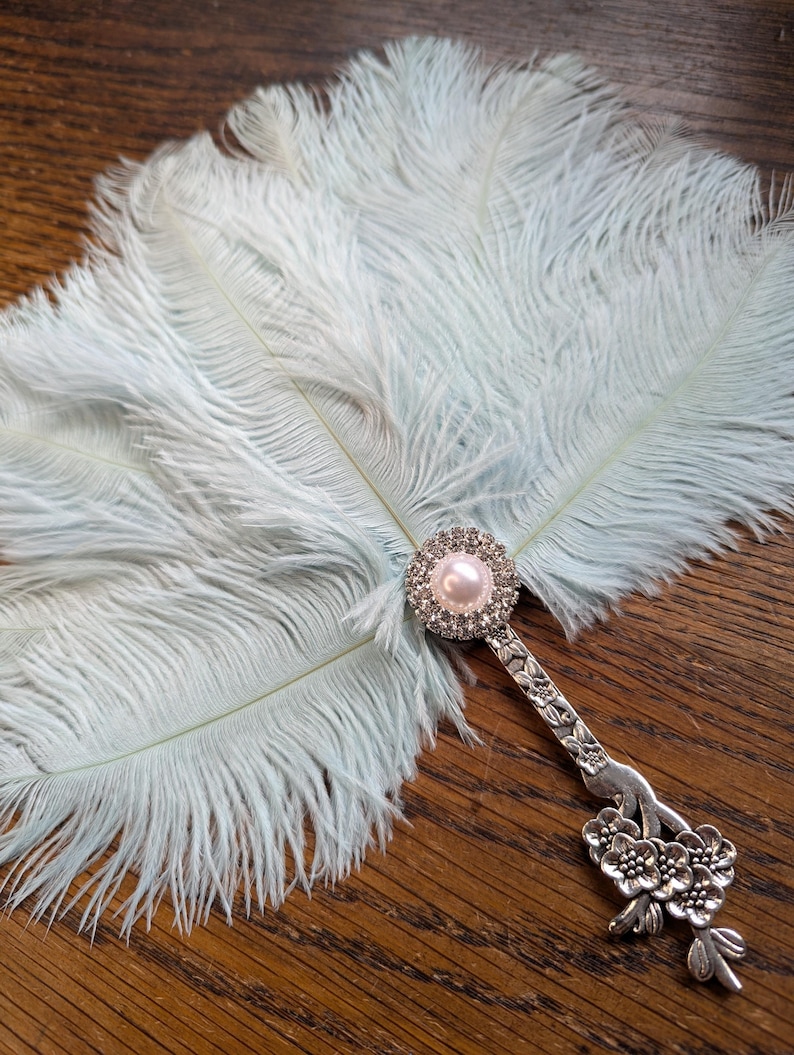 May include: A light blue feather fan with a decorative silver handle. The handle features a pearl and rhinestone embellishment at the top and a floral design at the base. The fan is made of soft, fluffy feathers.
