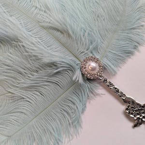 May include: A light blue ostrich feather fan with a decorative silver handle. The handle features a pearl and rhinestone embellishment at the base of the feathers and a floral design at the end.