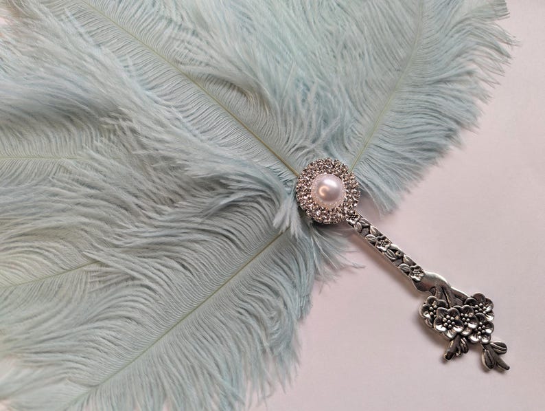 May include: A light blue ostrich feather fan with a decorative silver handle. The handle features a pearl and rhinestone embellishment at the base of the feathers and a floral design at the end.