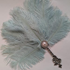 May include: A light blue feather fan with a decorative silver handle. The handle features a pearl and rhinestone embellishment. The fan is made of soft, fluffy feathers, perfect for a vintage or theatrical look.