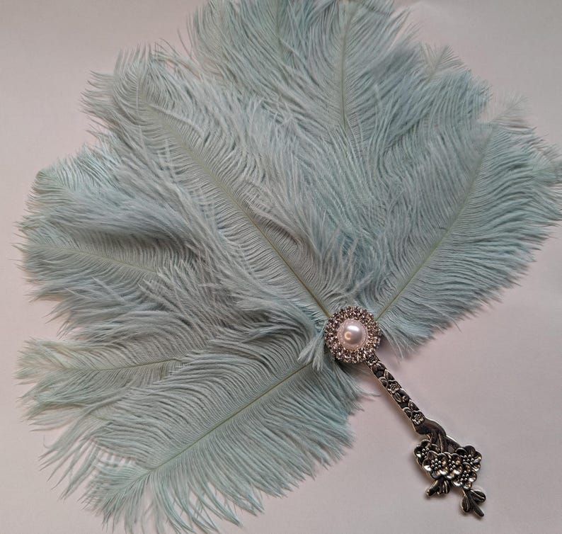 May include: A light blue feather fan with a decorative silver handle. The handle features a pearl and rhinestone embellishment. The fan is made of soft, fluffy feathers, perfect for a vintage or theatrical look.