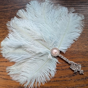 May include: A light blue feather fan with a silver handle. The fan features a central embellishment with a pink pearl surrounded by clear rhinestones. The handle is decorated with floral and leaf designs.