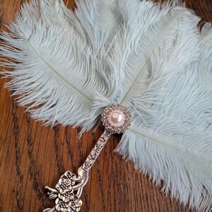 May include: A light blue feather fan with a decorative silver handle. The handle features a floral design and a pearl-centered, rhinestone-studded embellishment. The fan's feathers are soft and fluffy, creating a delicate and elegant accessory.