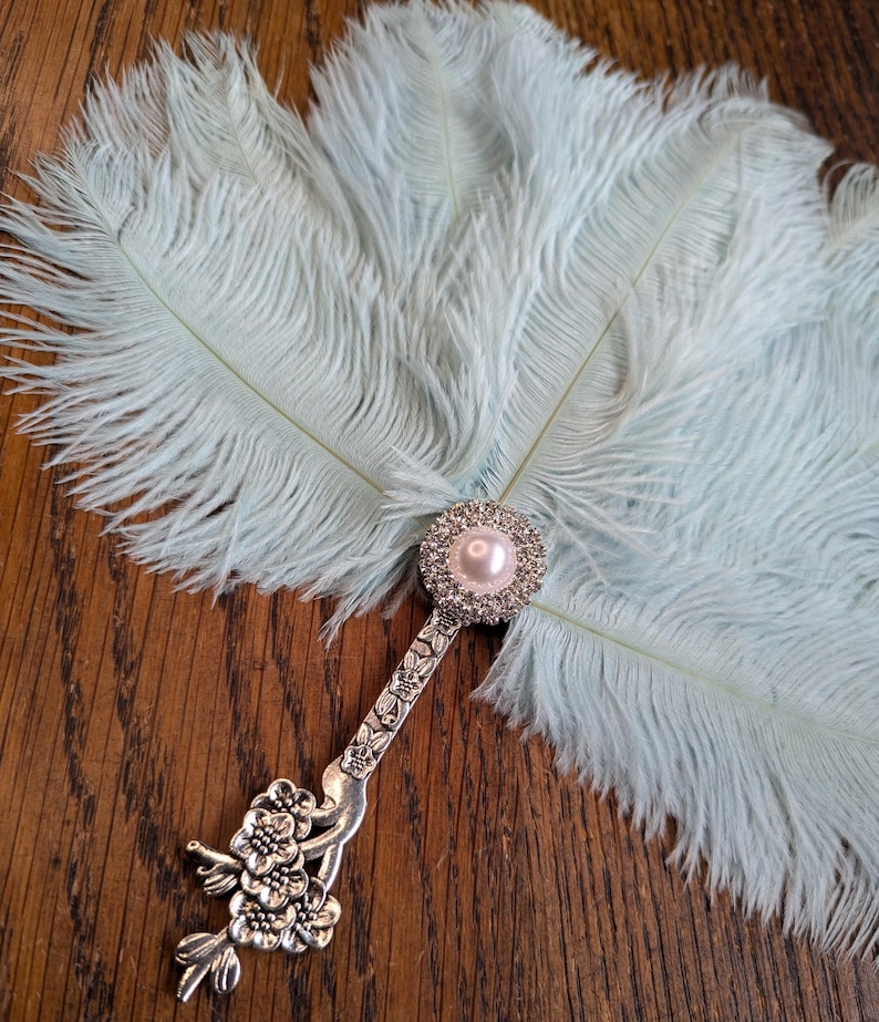 May include: A light blue feather fan with a decorative silver handle. The handle features a floral design and a pearl-centered, rhinestone-studded embellishment. The fan's feathers are soft and fluffy, creating a delicate and elegant accessory.
