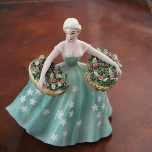 May include: A porcelain figurine of a woman in a teal dress with white flower accents, holding two baskets of roses. The figurine has blonde hair and is a decorative item.