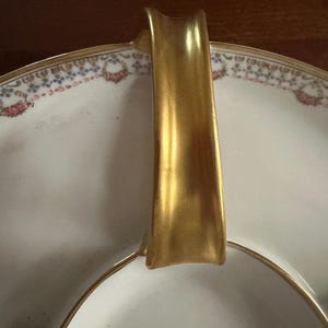 May include: A white porcelain serving dish with a gold handle and trim. The rim features a delicate floral pattern in pink, blue, and gold. The dish is divided into sections, ideal for serving appetizers or desserts.