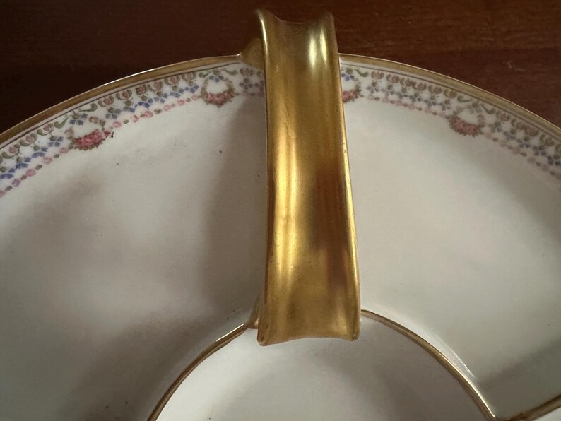 May include: A white porcelain serving dish with a gold handle and trim. The rim features a delicate floral pattern in pink, blue, and gold. The dish is divided into sections, ideal for serving appetizers or desserts.