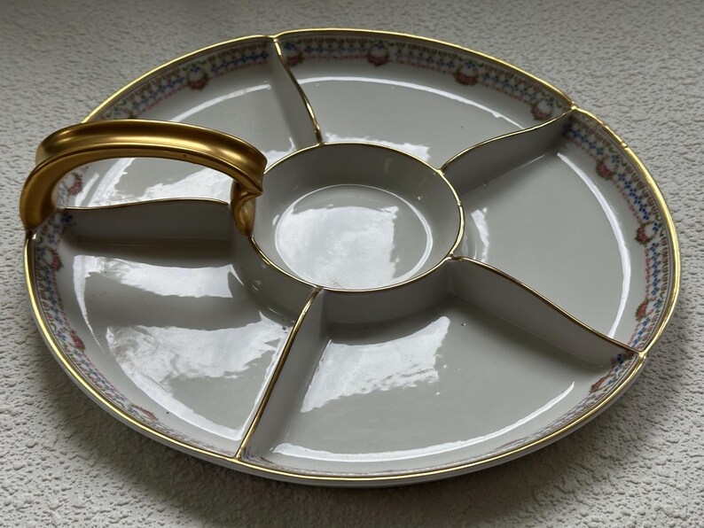 May include: A white divided serving platter with a gold handle and trim. The platter has a central bowl and five surrounding sections. The rim features a decorative pattern with blue, red, and gold accents.