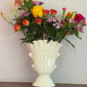 May include: A cream-colored ceramic vase with a scalloped rim and tiered shell-like design, filled with a vibrant bouquet of multicolored flowers including yellow roses, orange carnations, pink lilies, and purple filler blooms.