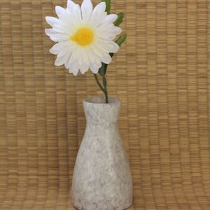 May include: A white ceramic vase with a textured surface holds a single white daisy with a yellow center. The vase and flower are on a brown woven mat.