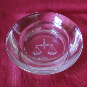 May include: A clear glass dish with a Libra zodiac symbol etched into the bottom. The symbol is a set of scales with the word "Libra" underneath.