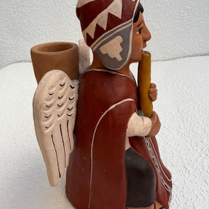 May include: A terracotta figurine of a kneeling figure wearing a red poncho and a patterned cap. The figure has white wings, holds a yellow flute, and features a cylindrical vessel on its back. The design includes dark brown accents and white line details.