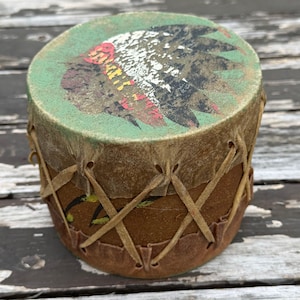 May include: A small cylindrical drum with a brown body and a green top surface featuring a weathered graphic of a feathered headdress. The drum is constructed with tan leather lacing that crisscrosses around the sides to secure the drumhead.