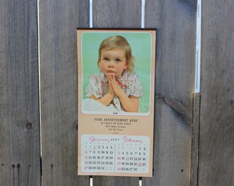 May include: A vintage 1997 calendar featuring a young girl with blonde hair and a white dress with pink flowers. The calendar has a light green background and the months of January and February are displayed.