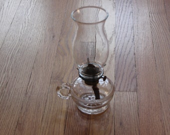 May include: A clear glass oil lamp with a round base and a tall, slender chimney. The lamp has a metal wick holder and a handle on the side.